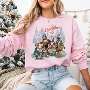 May include: A light pink sweatshirt featuring a graphic of Disney characters singing Christmas carols in a winter scene. The words "Merry Christmas" are written above the characters in a script font.