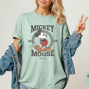 May include: A light green t-shirt featuring a distressed graphic of Mickey Mouse. The graphic includes the text "Mickey Mouse" and the year "1928" within a circular design. The shirt is part of the Comfort Colors Bay collection.