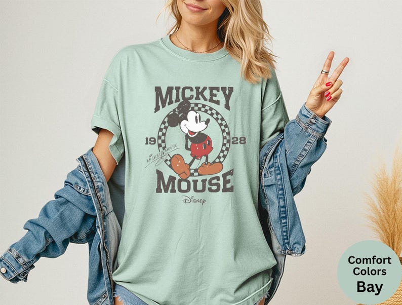 May include: A light green t-shirt featuring a distressed graphic of Mickey Mouse. The graphic includes the text "Mickey Mouse" and the year "1928" within a circular design. The shirt is part of the Comfort Colors Bay collection.