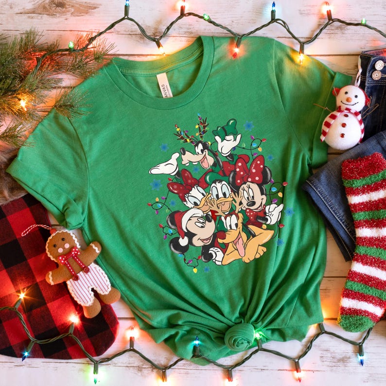 May include: A green t-shirt featuring a festive graphic of Disney characters, including Mickey and Minnie Mouse, Donald Duck, and Goofy, surrounded by Christmas lights. A snowman, gingerbread man, and Christmas stocking are also visible.