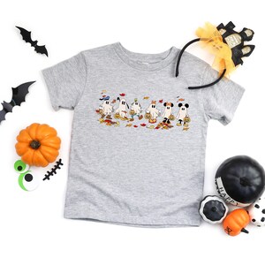 May include: A light gray t-shirt featuring a Halloween-themed graphic of cartoon ghosts holding jack-o'-lanterns. The shirt is surrounded by Halloween decorations, including pumpkins, bats, and a headband.