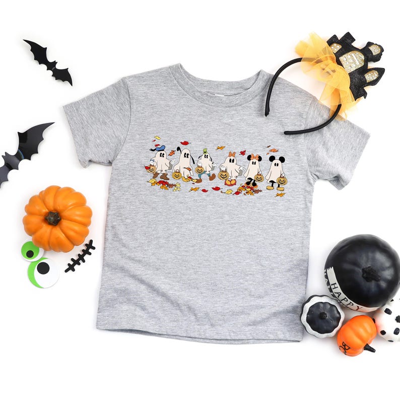 May include: A light gray t-shirt featuring a Halloween-themed graphic of cartoon ghosts holding jack-o'-lanterns. The shirt is surrounded by Halloween decorations, including pumpkins, bats, and a headband.
