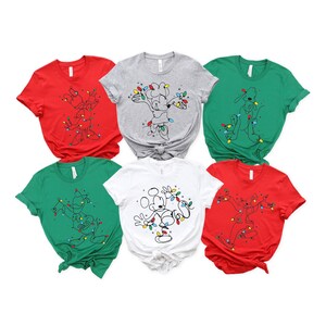 May include: A collection of six short-sleeved t-shirts in various colors: red, gray, green, and white. Each shirt features a cartoon character design wrapped in Christmas lights. The shirts are suitable for the holiday season.