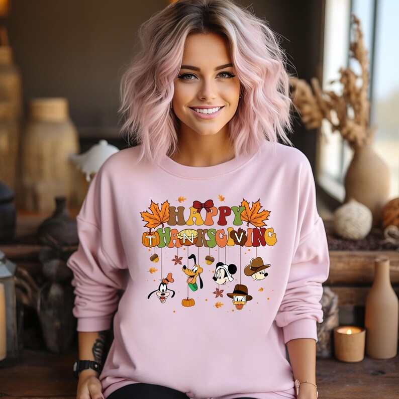 Op de afbeelding: Een lichtroze sweatshirt met de tekst "HAPPY THANKSGIVING" in kleurrijke letters en herfstthema graphics. Het ontwerp bevat cartoongezichten en herfstbladeren. Het sweatshirt wordt door een persoon gedragen.