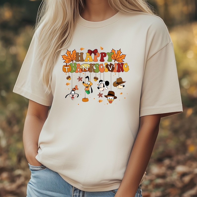 Op de afbeelding: Beige T-shirt met de tekst "HAPPY THANKSGIVING" in kleurrijke letters en herfstbladeren. Onder de tekst staan gezichten van stripfiguren met hoeden en strikken. Het shirt heeft een relaxte pasvorm.