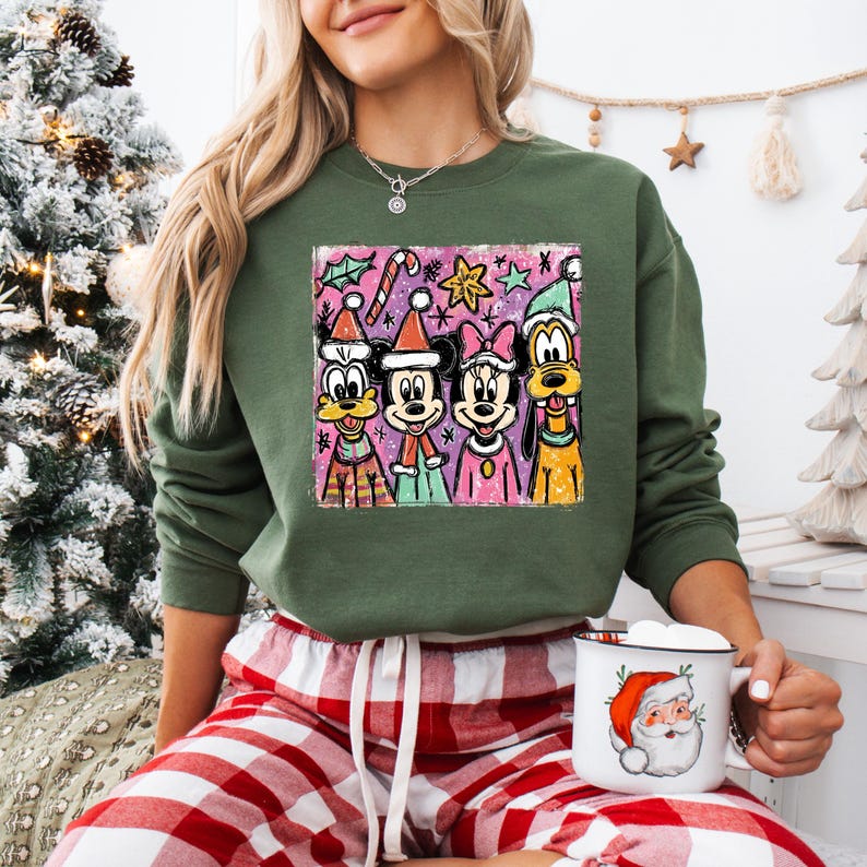 Op de afbeelding: Groene sweatshirt met een kleurrijke afbeelding van stripfiguren in kerstmutsen en feestelijke kleding. Het ontwerp omvat Mickey Mouse, Minnie Mouse, Donald Duck en Goofy. De persoon draagt ook een rood-witte geruite pyjamabroek en houdt een mok met een kerstmanontwerp vast.