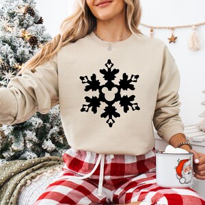 May include: A beige sweatshirt featuring a large black snowflake design with a Mickey Mouse silhouette in the center. The person is wearing red and white plaid pajama pants. A white mug with a Santa Claus graphic is held in the hand.