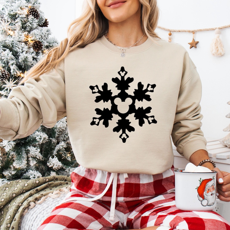 May include: A beige sweatshirt featuring a large black snowflake design with a Mickey Mouse silhouette in the center. The person is wearing red and white plaid pajama pants. A white mug with a Santa Claus graphic is held in the hand.