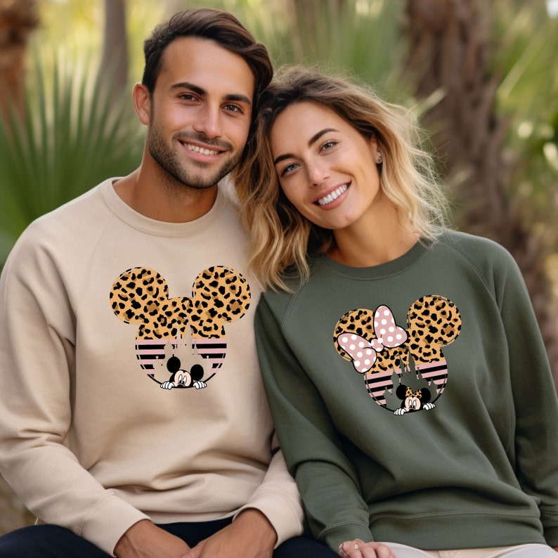 May include: A couple wearing matching sweatshirts with a cartoon image of Mickey Mouse and Minnie Mouse. The sweatshirts are beige and olive green with a leopard print design on the ears.
