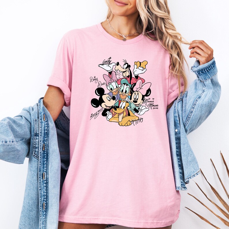 May include: Pink t-shirt featuring a graphic of Disney characters including Mickey Mouse, Minnie Mouse, Donald Duck, Daisy Duck, Goofy, and Pluto. The characters' names are written in a cursive font. The shirt has short sleeves.