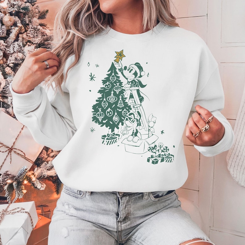 May include: White sweatshirt with a green illustration of Mickey Mouse and Pluto decorating a Christmas tree. Mickey is on a ladder placing a star on top of the tree. Pluto is sitting in a box with presents.