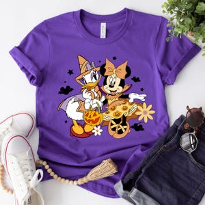 May include: Purple t-shirt with a graphic of Minnie Mouse and Daisy Duck dressed in Halloween costumes. Minnie Mouse is wearing a witch costume with a black hat and a pumpkin-themed dress. Daisy Duck is wearing a witch costume with a pointed hat and a star on her chest. They are both holding a pumpkin.