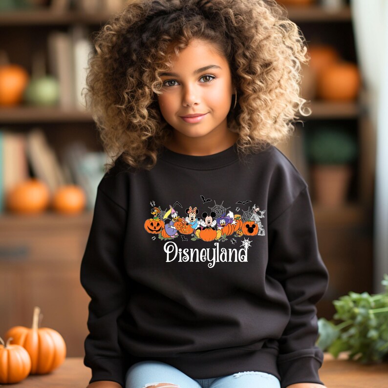K&ouml;nnte beinhalten: Schwarzes Sweatshirt mit einem Disneyland-Motiv mit Zeichentrickfiguren und K&uuml;rbissen. Das Wort "Disneyland" steht in wei&szlig;er Schrift darunter. Getragen von einer jungen Person.