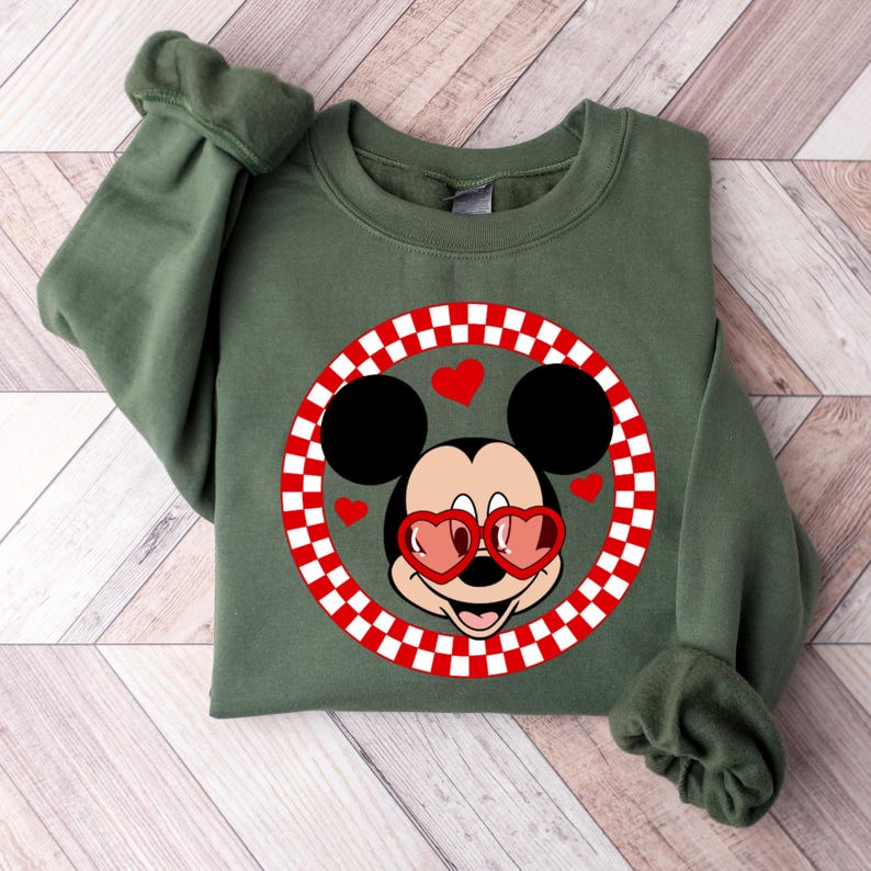 May include: A green sweatshirt with a red and white checkered circle featuring Mickey Mouse wearing heart-shaped sunglasses.  Mickey Mouse is smiling and has red hearts around his head.