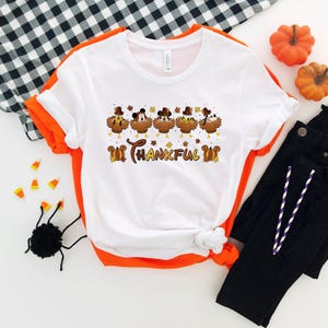 May include: White t-shirt with an orange trim featuring a Thanksgiving-themed graphic. The graphic includes cartoon turkeys wearing hats and the word "THANKFUL". The shirt is paired with black pants, pumpkins, and Halloween decorations.