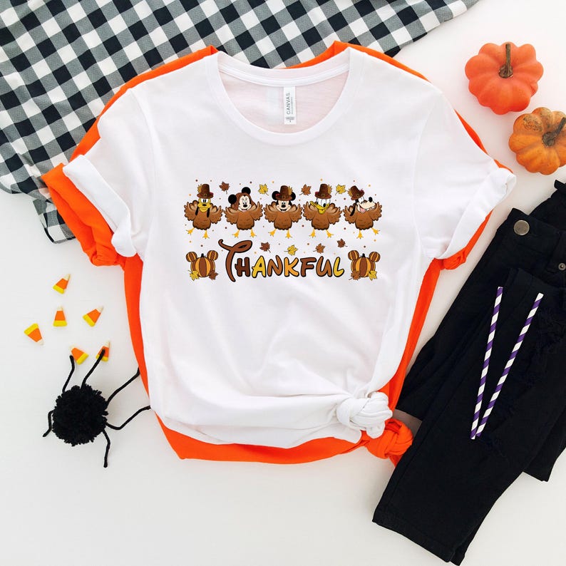 May include: White t-shirt with an orange trim featuring a Thanksgiving-themed graphic. The graphic includes cartoon turkeys wearing hats and the word "THANKFUL". The shirt is paired with black pants, pumpkins, and Halloween decorations.