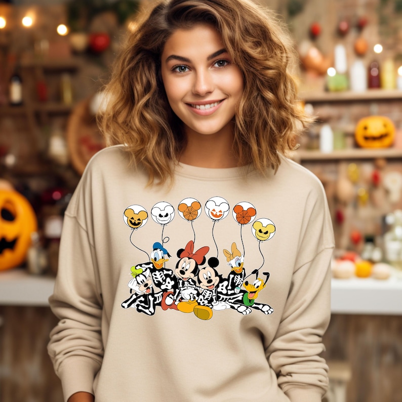 May include: A tan sweatshirt with a graphic of Mickey Mouse, Minnie Mouse, Donald Duck, Goofy, and Pluto dressed as skeletons holding Halloween-themed balloons. The balloons are orange, white, and black with jack-o'-lantern faces.