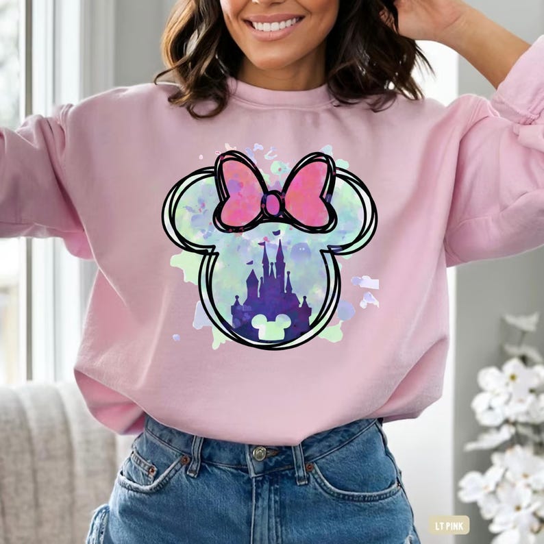 May include: A pink sweatshirt with a watercolor design of Mickey Mouse ears with a pink bow and a silhouette of Cinderella's castle inside.