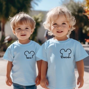 Puede incluir: Dos ni&ntilde;os peque&ntilde;os con camisetas azules claras con un gr&aacute;fico de cabeza de Mickey Mouse negro. La camiseta de la izquierda dice "Ben" y la camiseta de la derecha dice "Brother".