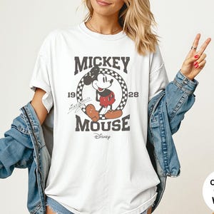 May include: White t-shirt featuring a distressed graphic of Mickey Mouse. The design includes the text "Mickey Mouse" and the years "1928" and "19". The shirt has a classic crew neck and short sleeves. The shirt is a comfort color.