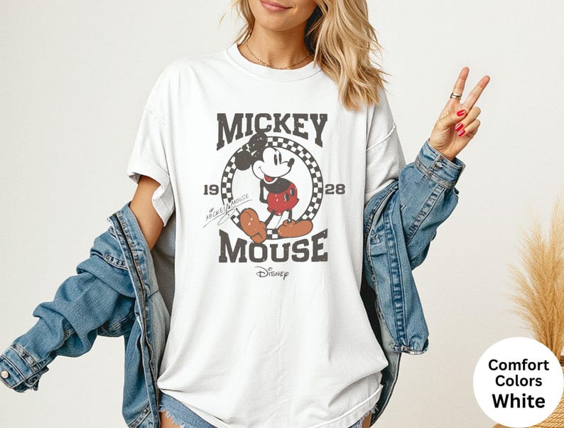May include: White t-shirt featuring a distressed graphic of Mickey Mouse. The design includes the text "Mickey Mouse" and the years "1928" and "19". The shirt has a classic crew neck and short sleeves. The shirt is a comfort color.