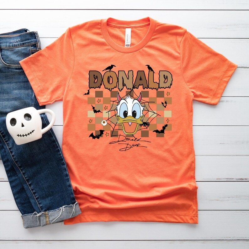 May include: Orange t-shirt featuring a Halloween-themed Donald Duck graphic. The design includes the word "DONALD" in brown letters, bats, spiders, and a signature. The shirt is paired with blue jeans and a skull-shaped mug.