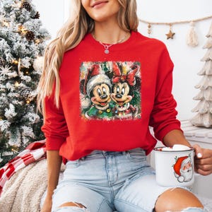 May include: A red sweatshirt featuring a festive graphic of Mickey and Minnie Mouse in Christmas attire. The graphic shows Mickey and Minnie wearing Santa hats and holiday outfits. The sweatshirt is a long-sleeved crew neck.