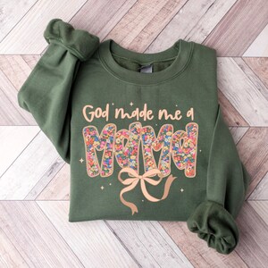 Pu&ograve; includere: Una felpa verde oliva con la scritta "God made me a Mama" in un design floreale. Un fiocco marrone chiaro &egrave; sotto la parola "Mama". La felpa &egrave; piegata su una superficie di legno marrone chiaro.