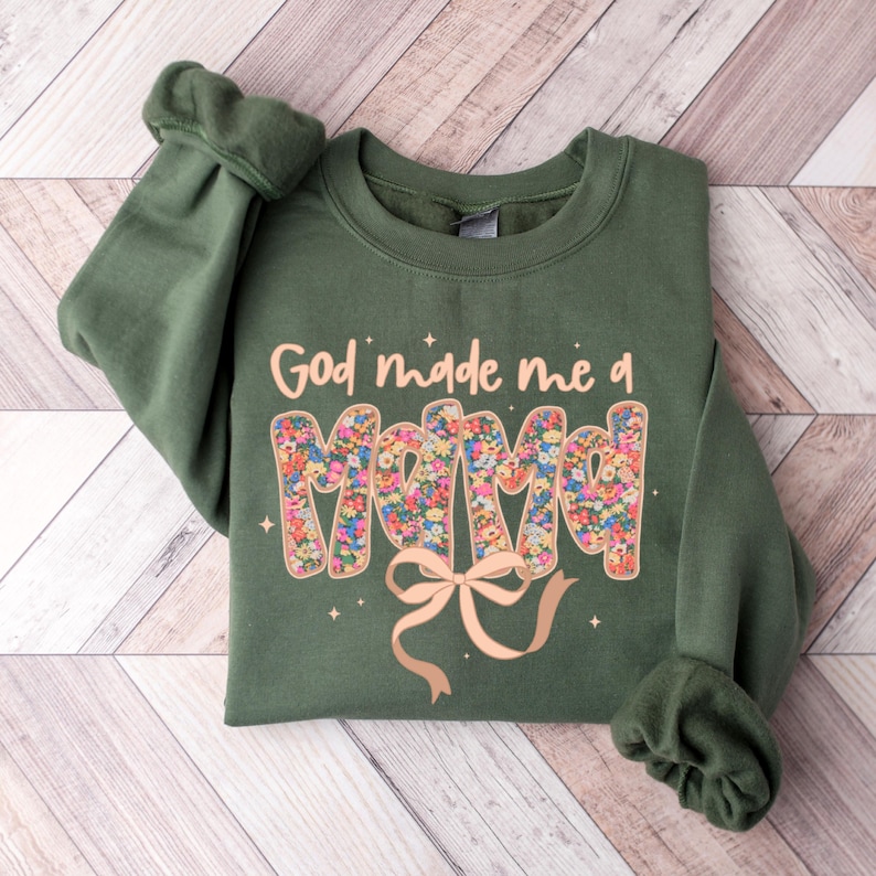 Pu&ograve; includere: Una felpa verde oliva con la scritta "God made me a Mama" in un design floreale. Un fiocco marrone chiaro &egrave; sotto la parola "Mama". La felpa &egrave; piegata su una superficie di legno marrone chiaro.