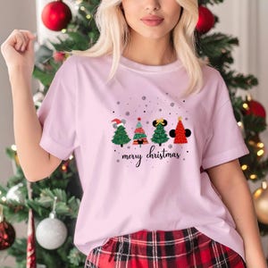 May include: A light pink t-shirt featuring four Christmas tree designs with mouse ears and a "merry christmas" script. The trees are green, red, and black with snowflake accents. The shirt is made of a soft material.