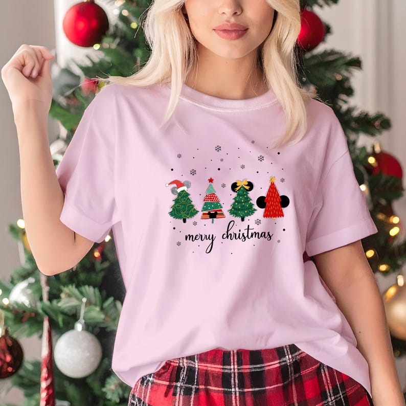 May include: A light pink t-shirt featuring four Christmas tree designs with mouse ears and a "merry christmas" script. The trees are green, red, and black with snowflake accents. The shirt is made of a soft material.
