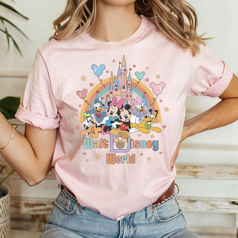 Puede incluir: Camiseta rosa con un gr&aacute;fico de Mickey Mouse y sus amigos frente al Castillo de Cenicienta en Walt Disney World. El gr&aacute;fico tambi&eacute;n incluye un arco&iacute;ris y estrellas.