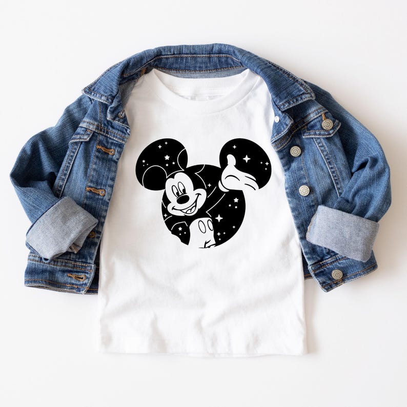 May include: White t-shirt with a black and white Mickey Mouse design. The graphic shows Mickey's face and ears filled with stars. A blue denim jacket is draped over the shirt.