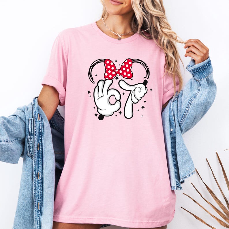 K&ouml;nnte beinhalten: Hellrosa T-Shirt mit einem Minnie Mouse-inspirierten Design. Das Design umfasst eine rote Schleife mit wei&szlig;en Punkten, wei&szlig;e Handschuhe, die die Zahl 67 bilden, und schwarze Akzente. Ein l&auml;ssiges Kleidungsst&uuml;ck f&uuml;r den Alltag.