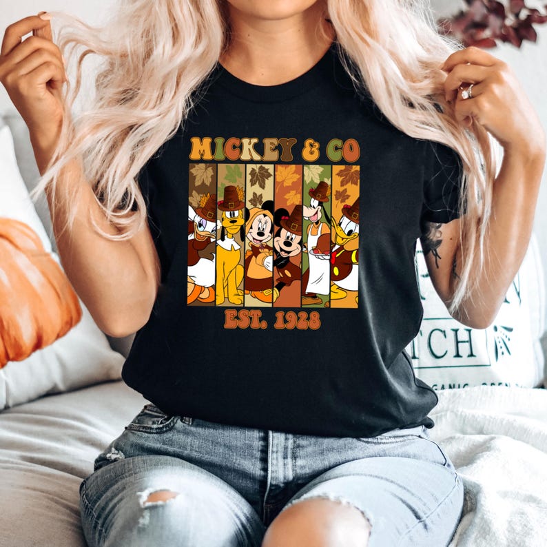 May include: Black t-shirt featuring a graphic of Mickey & Co. characters in fall attire. The design includes Donald Duck, Pluto, Mickey Mouse, Goofy, and others, with the text "MICKEY & CO" and "EST. 1928" in a retro font.