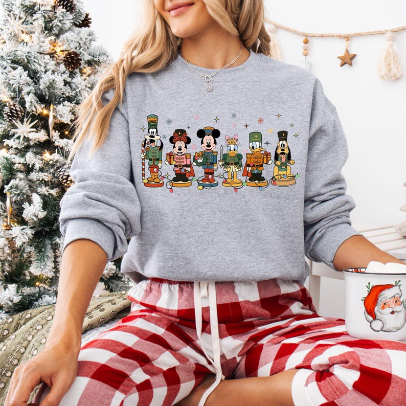 May include: A gray sweatshirt featuring a colorful graphic of Disney characters dressed as nutcrackers. The characters are arranged in a row, each with unique uniforms and poses. The sweatshirt is paired with red and white plaid pajama pants, creating a festive holiday theme.