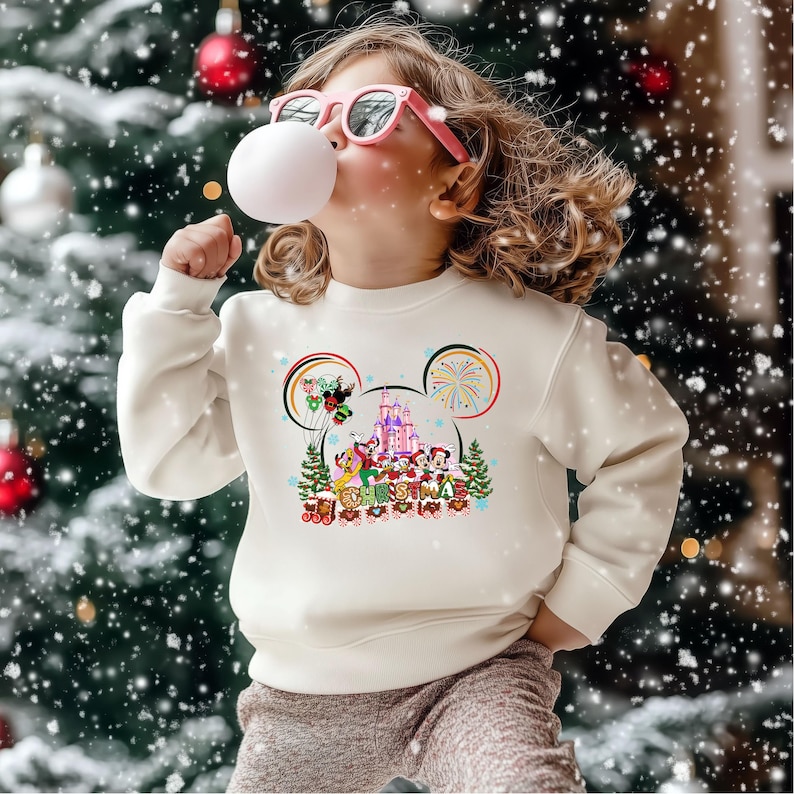May include: A cream-colored sweatshirt featuring a colorful Disney Christmas design. The graphic includes Mickey Mouse ears, a castle, and various Disney characters. The sweatshirt is worn by a child wearing pink sunglasses, blowing a bubble.