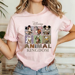 May include: Pink t-shirt with a graphic of Disney characters in safari outfits. The characters are Mickey Mouse, Donald Duck, Goofy, and Pluto. The text "Disney Animal Kingdom" is printed below the graphic.