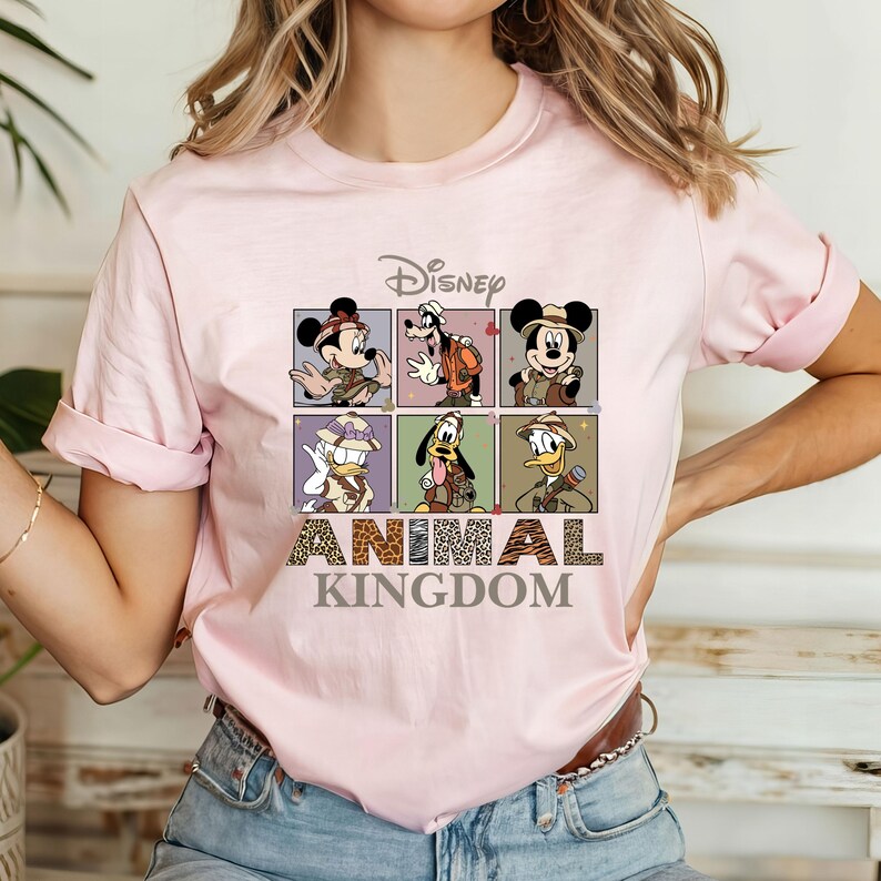 May include: Pink t-shirt with a graphic of Disney characters in safari outfits. The characters are Mickey Mouse, Donald Duck, Goofy, and Pluto. The text "Disney Animal Kingdom" is printed below the graphic.