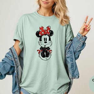 May include: A light green t-shirt featuring a black and white Minnie Mouse graphic with a red polka dot bow. The shirt is part of the Comfort Colors Bay collection and is paired with a denim jacket.
