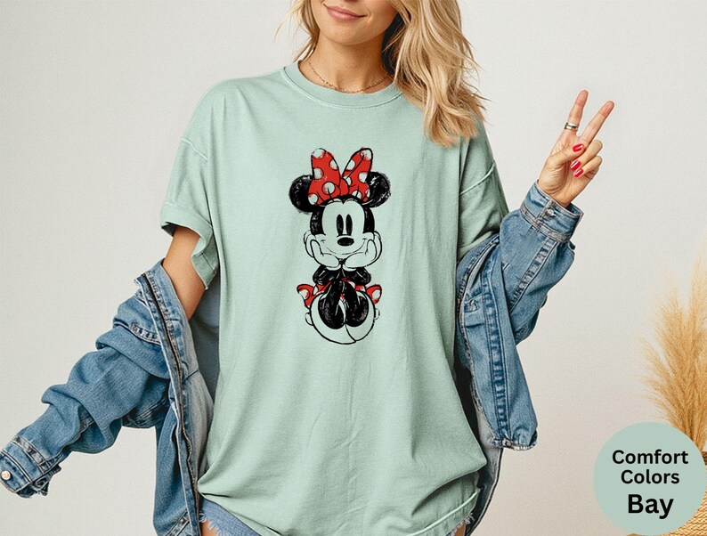 May include: A light green t-shirt featuring a black and white Minnie Mouse graphic with a red polka dot bow. The shirt is part of the Comfort Colors Bay collection and is paired with a denim jacket.