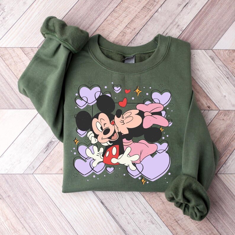 May include: A green sweatshirt with a graphic print of Mickey Mouse and Minnie Mouse kissing. The graphic is surrounded by purple hearts and stars.