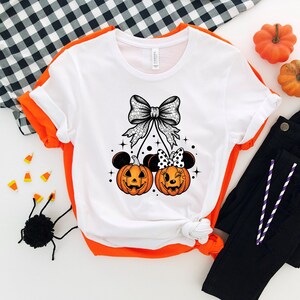 May include: White t-shirt with a Halloween-themed design. The graphic features two orange jack-o'-lanterns with mouse ears, a large bow, and stars. Black pants and pumpkins are also visible.