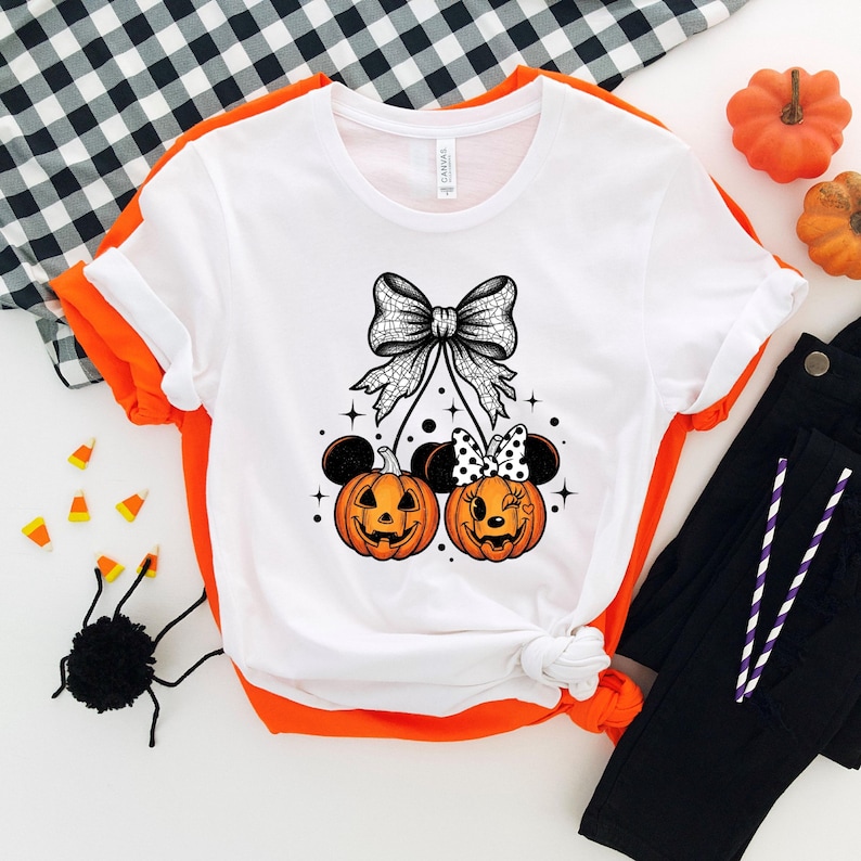 May include: White t-shirt with a Halloween-themed design. The graphic features two orange jack-o'-lanterns with mouse ears, a large bow, and stars. Black pants and pumpkins are also visible.