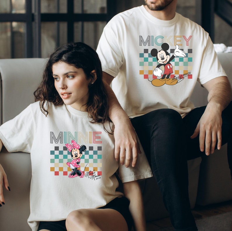 May include: A couple wearing matching white t-shirts with a retro checkered pattern and cartoon images of Mickey and Minnie Mouse. The shirts feature the text "Mickey Mouse" and "Minnie Mouse" in a colorful, retro font.