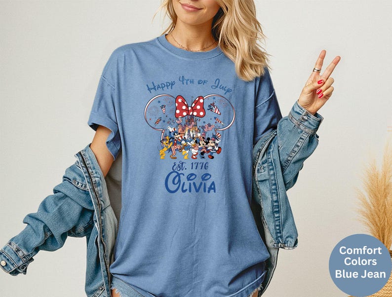 Pode incluir: Uma camiseta azul Comfort Colors com um gr&aacute;fico celebrando o 4 de julho. O design inclui orelhas do Mickey Mouse, um la&ccedil;o vermelho e v&aacute;rios personagens da Disney. O texto "Happy 4th of July" e "Olivia" tamb&eacute;m est&atilde;o presentes.