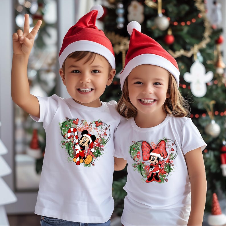 May include: Two children wearing white t-shirts with Christmas-themed Mickey Mouse and Minnie Mouse graphics. The children are wearing red Santa hats.