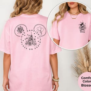 May include: A light pink t-shirt with a graphic of a castle, fireworks, and balloons inside a Mickey Mouse head shape. The text "THE HAPPIEST PLACE ON EARTH" is printed around the design. A smaller graphic of Mickey Mouse is on the front.