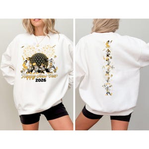 May include: White sweatshirt with a festive design for the New Year 2026. The front features a gold and black graphic with fireworks and cartoon characters. The back has a vertical line of cartoon characters in celebratory poses.