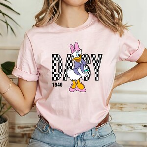 May include: Pink t-shirt with a cartoon image of Daisy Duck wearing a purple dress and a pink bow. The text "Daisy 1940" is printed below the image.